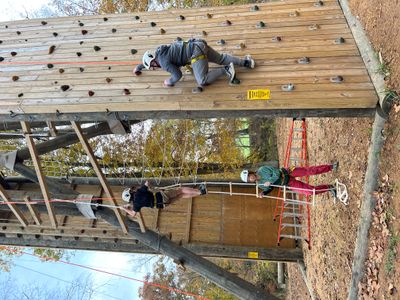 On the Tango Tower climbing structure, one student climbs a typical rock wall, while another student climbs a giant wooden ladder and a teacher helps steady a rope ladder.
