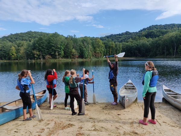 During a paddling lesson at SAS, a SAS instructor shows a paddle to a small group of students. All are wearing life jackets and carrying paddles, standing on a sandy beach at the shore of a lake under blue sky.