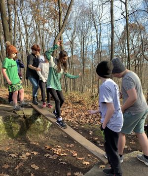 A group of students stands on boulders in a forest, working together to build a bridge out of planks of wood.