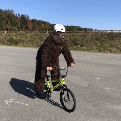 A person wearing a sasquatch suit and a bike helmet rides a BMX bike, coasting across a concrete pad while standing on the pedals.
