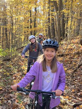 Students on mountain bikes smile into the camera, while riding a flat, forested trail surrounded by autumn leaves.