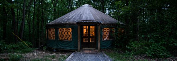 A round, cozy yurt is lit up from inside, surrounded by forest at dusk.