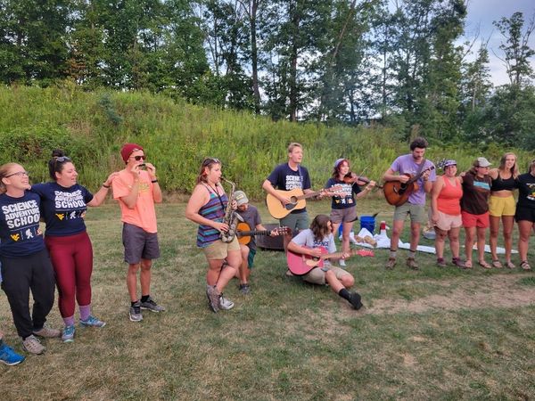 SAS staff sing Country Roads around a campfire, arms around each others' shoulders. Several staff play musical instruments: guitars, a saxophone, a fiddle, and a harmonica.