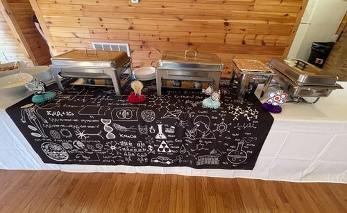A buffet table is laid for dinner in a wooden building. The tablecloth is patterned with science imagery, including a DNA molecule, various chemical symbols, and a diagram of the human body.