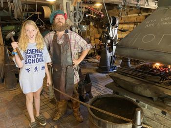 Inside a blacksmith's shop full of metal tools, a smiling student wearing a Science Adventure School t-shirt holds a large hammer, while standing next to a blacksmith and a forge with visible fire.