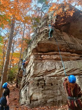 Two students climb outdoors on a natural rock face, while belayers and other students watch.
