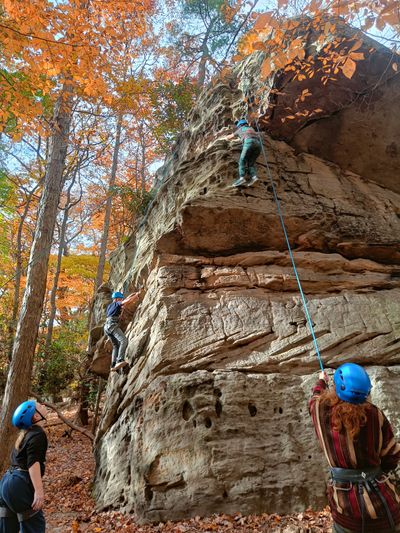 Two students wearing harnesses and helmets rock climb on a very large boulder, while staff belay and other students watch from the ground.