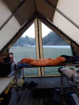 The inside of a canvas tent at the Summit: two bunk beds are visible, with sleeping bags and pillows. The screen door of the tent is open, showing a grassy field.