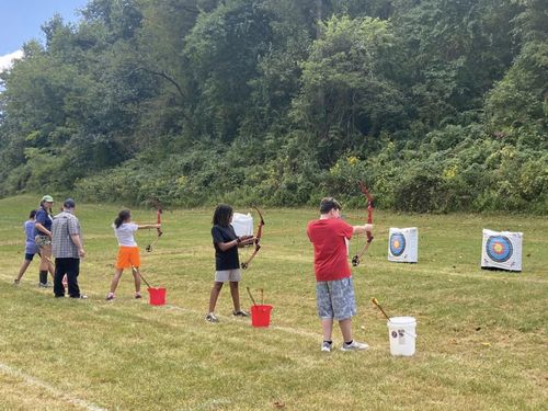 Students in archery class shoot colorful compound bows at targets, in a grassy field.