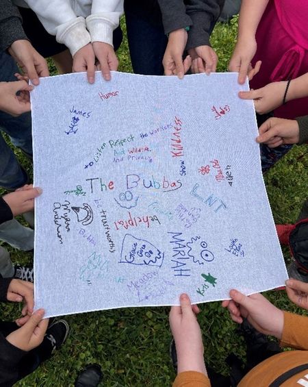 Taken from above, this photo shows students' hands holding a square, white bandana, on which they have signed their names and written words and phrases like "be helpful," "be trustworthy," and "respect the workers and wildlife and privacy."