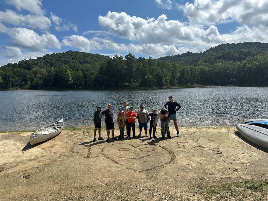Students pose after a paddling lesson on a sandy lakeside beach, where they have written "I Heart SAS" and a smiley face into the sand with a stick.