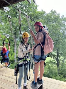 A smiling girl wearing a helmet and harness stands on a wooden platform at the top of a zip line, while a woman connects a zip trolley to the girl's harness.