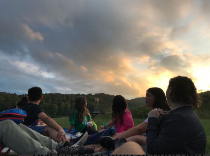 Students seated on a grassy field watch the sky, which is blue, gold, and orange with dramatic gray clouds.