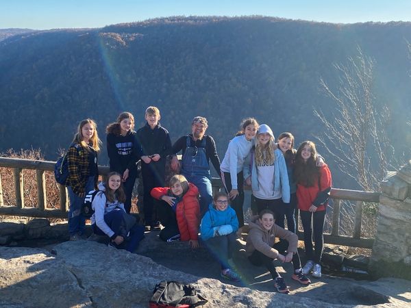Students and their teacher pose for a group photo at an overlook, with a dramatic forested canyon and a blue sky behind them.