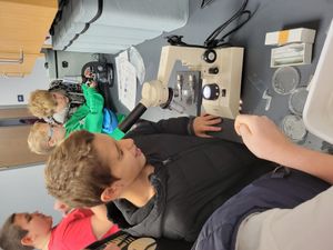 Students peer into microscopes in an indoor lab. Petri dishes, slides, and identification keys are on the table near the microscopes.