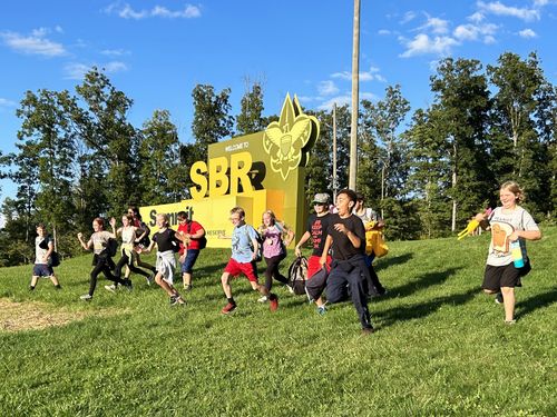 A dozen students laugh and run down a hill in front of a large sign that says Welcome to SBR.
