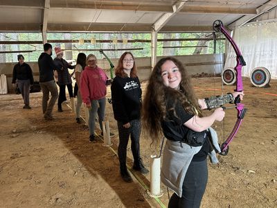 Smiling students line up with bows during an archery class in an indoor range.