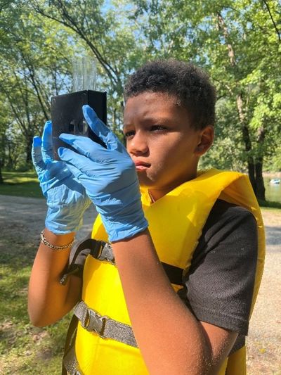 A student stands outdoors surrounded by trees, wearing a yellow life jacket and blue rubber gloves examines a piece of scientific equipment.