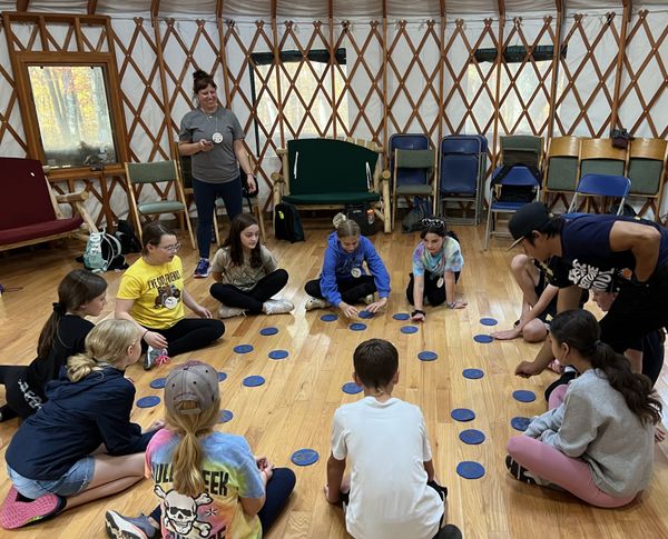 Students play a game inside a yurt, seated on the floor with colorful plastic spots on the floor between them.