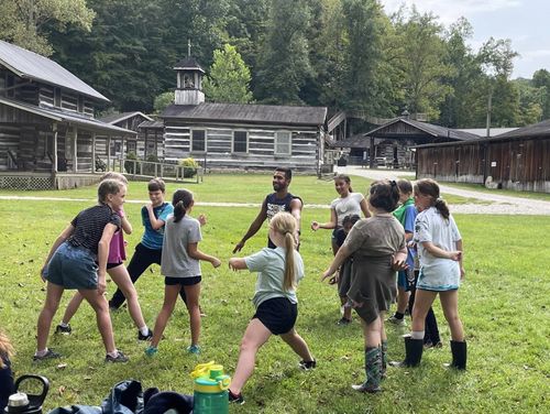 A group of students plays a group game in front of historic buildings at Heritage Farm.