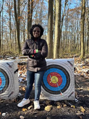 A student smiles proudly while posing next to an archery target. Three arrows have been shot, two of which were bulls-eyes.