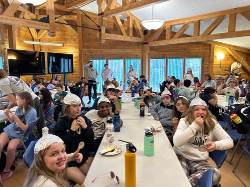 Students wearing silly tinfoil hats sit at long tables for dinner, in a large room with wood walls and rafters and large windows.