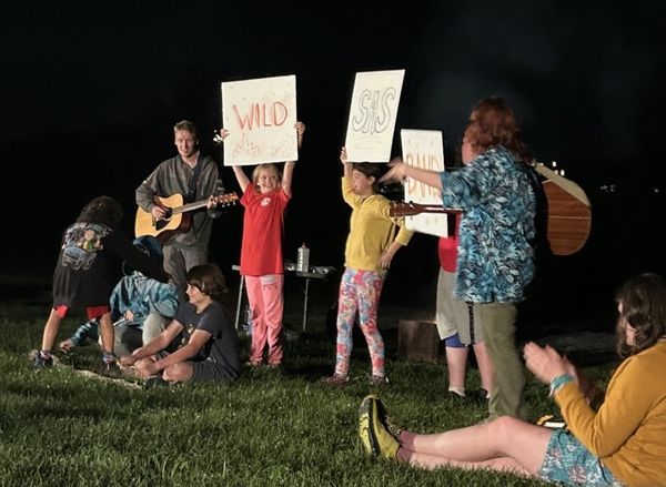 A cheerful campfire scene: Outdoors at night, three students stand, holding large signs saying "WILD SAS BAND," next to two SAS staff holding guitars. Other students are seated nearby. 