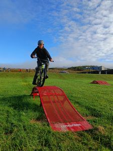 A boy with a determined expression rides a BMX bike over a bright red metal ramp in a grassy field, while his classmates watch from a distance.