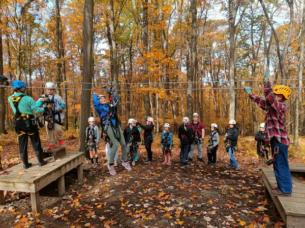Students practice braking on a small zip line, surrounded by a forest in bright fall color.