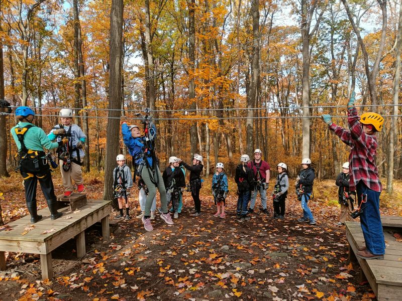 Students wearing helmets and harnesses learn to zip line on a ground school, a small model zip line. Two staff members coach one student across the zip, while other classmates watch.