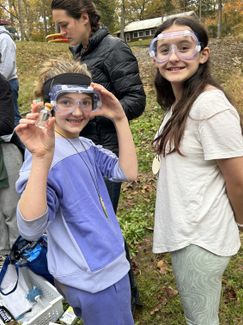 Two students wear safety goggles while performing an outdoor science experiment. One student holds up a small glass flask.