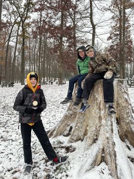 Three students smile in a forest lightly dusted with snow. Two students are sitting on a giant tree stump.